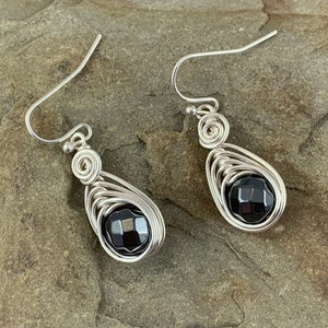 May include: A pair of silver wire wrapped earrings with black gemstone accents. The earrings are teardrop shaped and feature a delicate wire design.