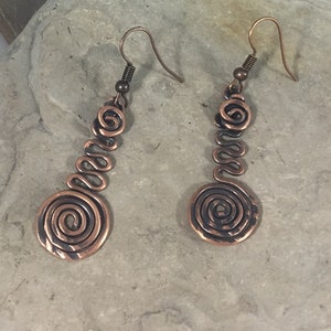 May include: A pair of long, copper earrings with a spiral design. The earrings are made of wire that is twisted into a series of spirals, with a larger spiral at the bottom. The earrings are a warm copper color.