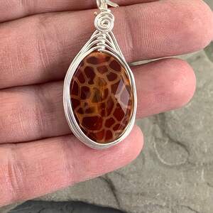 May include: A silver wire wrapped pendant featuring a brown and orange agate stone with a unique pattern. The pendant is oval shaped and hangs from a silver chain.