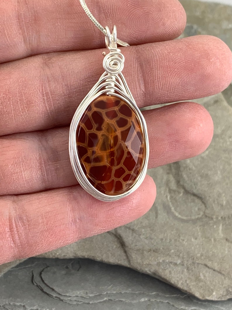 May include: A silver wire wrapped pendant featuring a brown and orange agate stone with a unique pattern. The pendant is oval shaped and hangs from a silver chain.