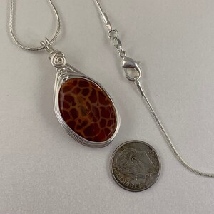May include: A silver chain necklace with a brown and white agate stone pendant wrapped in silver wire. The pendant is about the size of a US quarter.
