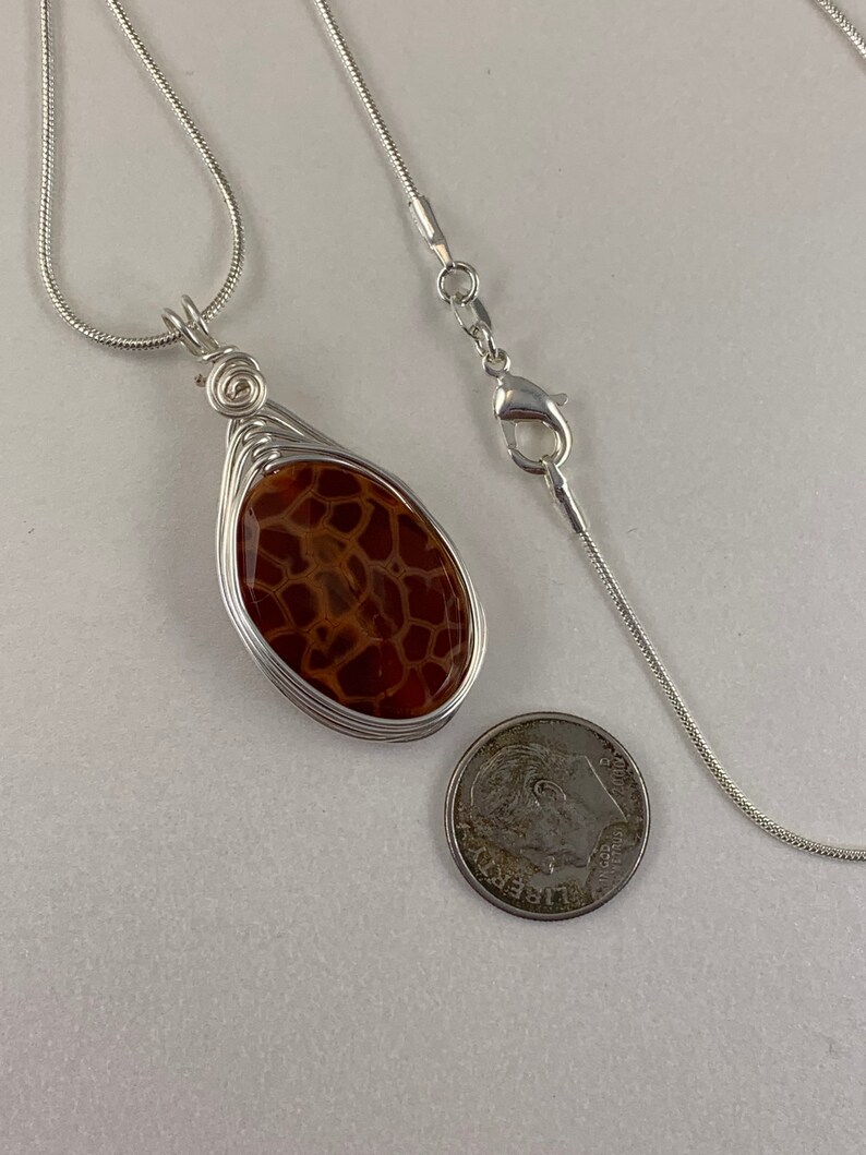 May include: A silver chain necklace with a brown and white agate stone pendant wrapped in silver wire. The pendant is about the size of a US quarter.