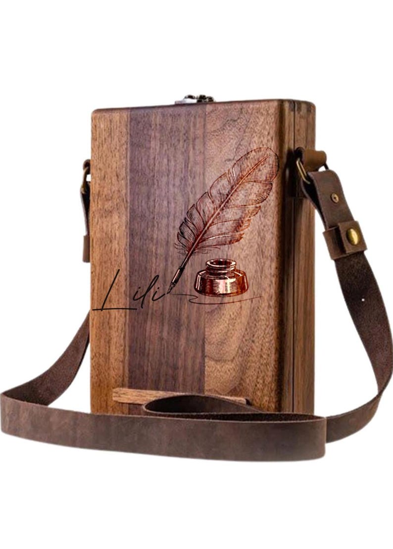May include: A brown wooden box with a leather strap. The box has a design of a feather and inkwell with the word "Lili" written below it.