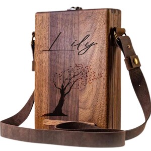 May include: A personalized wooden box with a brown leather strap. The box has a tree design with leaves blowing in the wind and the name "Lily" engraved on the front.