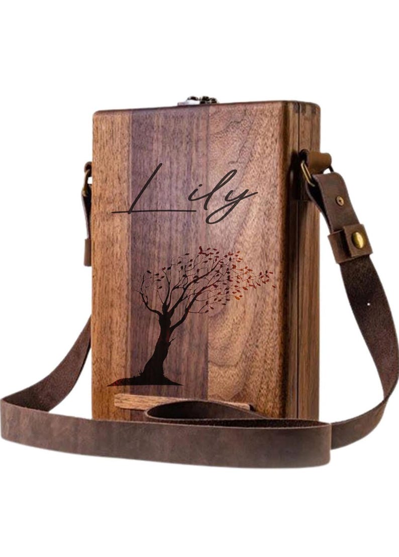 May include: A personalized wooden box with a brown leather strap. The box has a tree design with leaves blowing in the wind and the name "Lily" engraved on the front.