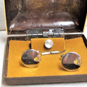 May include: Vintage cufflinks and tie clip set in a brown and gold box. The cufflinks are silver-toned with a curved design, and the tie clip is a matching silver tone. The box has a gold interior and a hinged lid.