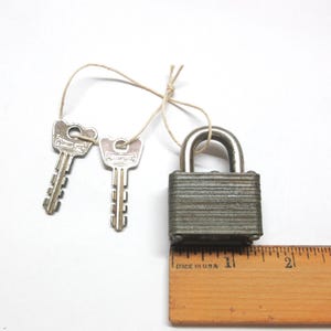 May include: A vintage metal padlock with a silver shackle and a textured, gray body. Two matching keys are attached with a string. A wooden ruler with "MADE IN USA" is in the foreground.
