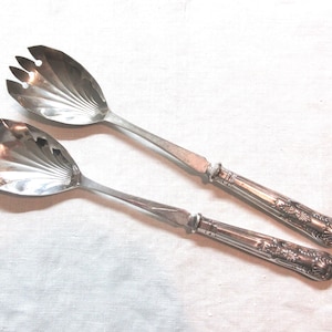 May include: A pair of vintage silver salad servers. One server has a spoon-shaped bowl, the other has a fork-like design. Both have ornate handles with floral patterns and are set against a white background.