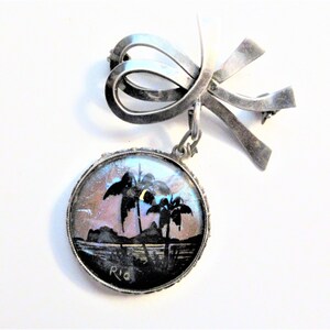 May include: A vintage silver-toned brooch with a bow design and a dangling round pendant. The pendant features a painted landscape of palm trees and the word "Rio". The brooch is a unique piece of jewelry.
