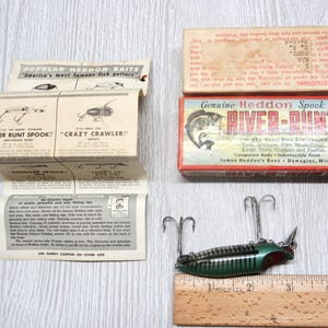 May include: A vintage Heddon River-Runt fishing lure in a green and black striped finish with two metal treble hooks, accompanied by its original cardboard box and a folded paper catalog insert, all positioned next to a wooden ruler.