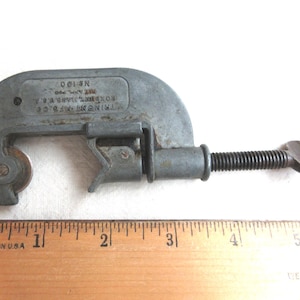 May include: A vintage gray pipe cutter with a blade and a threaded adjustment screw. The tool has the text "TRIMO MFG. CO. ROXBURY, MASS. U.S.A." and "No. 100" stamped on it. A wooden ruler with "MADE IN U.S.A." is next to the cutter.