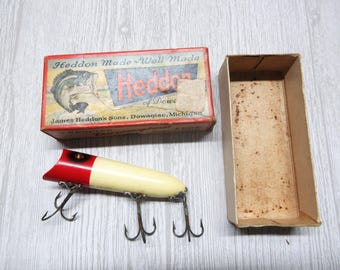 May include: A vintage fishing lure with a cream-colored body and a red head, featuring three metal treble hooks. Beside it is a rectangular cardboard box labeled Heddon Made Well Made, featuring a fish illustration and text.