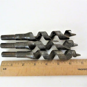 May include: Three metal drill bits with a twist design, laid out on a wooden ruler. The ruler is marked with inches and the text "MADE IN USA" and "G-H".