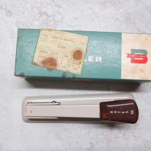May include: An overhead shot of a vintage stapler and its box. The stapler is beige with a brown handle, and the box is teal and white with the word "Bates" and a logo. A small, aged paper is on the box.