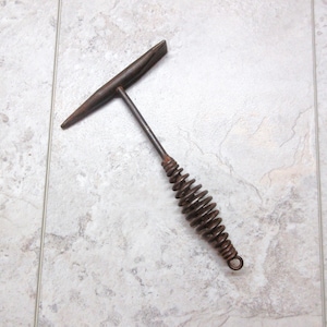 May include: A vintage metal welding hammer with a T-shaped head and a coil spring handle. The hammer is made of dark brown metal and appears to be well-used. The tool is designed for welding and metalworking.