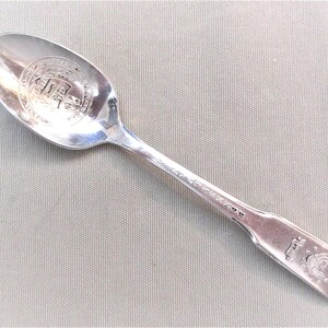 May include: A silver spoon with a detailed design on the bowl and handle. The bowl features a circular emblem with text and a building. The handle has a decorative pattern and an emblem near the end.