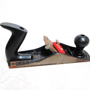 May include: A vintage hand plane with a black handle and knob. The plane has a metal body with a blade and a red and brown wooden piece. The tool is designed for woodworking and has a classic, well-used appearance.