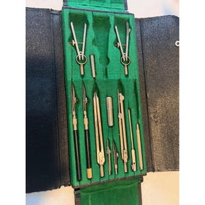 May include: A vintage drafting instrument set housed in a black folding case with a green felt-lined interior. The set includes various metal compasses, dividers, ruling pens with black handles, and small cylindrical metal attachments for precision work.