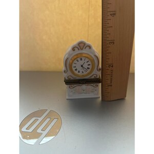 May include: A white porcelain clock-shaped trinket box with gold trim, a clock face featuring Roman numerals, and pink floral accents. A wooden ruler placed next to the object indicates a height of approximately 4 inches for the clock.