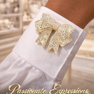 May include: A gold-toned bow-shaped cufflink, encrusted with sparkling crystals, adorns a white dress shirt cuff. The jewelry piece is a statement accessory. The image also includes the text "Passionate Expressions Jewelry by: Kelly Boyd."