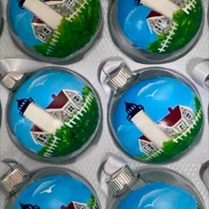 May include: Six glass ornaments with a painted design of a lighthouse with a white tower, red roof, and a white picket fence. The ornaments are painted on a blue background with white clouds and a seagull.