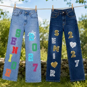 May include: Two pairs of denim jeans hanging on a clothesline. The light blue pair features the word SENIOR in colorful block letters and the number 2027 in blue. The dark blue pair features the word SENIOR and 2027 in sparkling rhinestone letters.