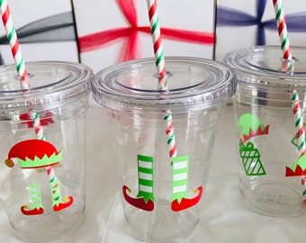 May include: Three clear plastic cups with lids and straws. Each cup has a different Christmas elf design. One cup has an elf with green and white striped legs, one has an elf with a red hat and green and white striped legs, and one has an elf with a red hat and a gift.