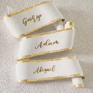 May include: Three white ribbons with gold trim and gold lettering. The names "George", "Adam", and "Abigail" are written on the ribbons. The ribbons are curled and arranged in a decorative manner.