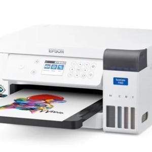 Może przedstawiać: Biała drukarka sublimacyjna Epson SureColor F100 z małym cyfrowym wyświetlaczem, przyciskami sterującymi i zintegrowanym systemem zbiorników z tuszem po prawej stronie. Z drukarki wysuwa się arkusz papieru z kolorową, abstrakcyjną grafiką.