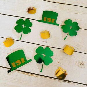 May include: St. Patrick's Day-themed stickers featuring green shamrocks, green hats with gold text, and gold pots of gold. The stickers are on a white wooden surface.