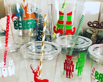 May include: Set of clear plastic cups with festive Christmas designs. The designs include a reindeer, a nutcracker, an elf, a cupcake, and the word "Believe".