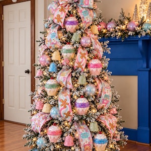 May include: A flocked Christmas tree decorated with pastel-colored cupcake ornaments, small bell-shaped ornaments, and iridescent baubles. The tree is wrapped in wide ribbons featuring gingerbread men and tea cup patterns, topped with a large bow.