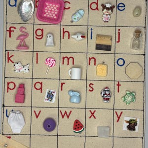 May include: A grid of small decorative items, each in a separate square, including a seashell, a pink basket, a baby shoe, a chef figurine, a pink flamingo, a ghost, a small airplane, a ruler, a small jar, a cat, a lollipop, a white mug, a sponge, a wooden shape, a stack of blocks, a playing card, a blue bunny, a Santa figurine, a turtle, a pair of underwear, a purple ball, a watermelon slice, a clear cube, and a Grogu figurine.