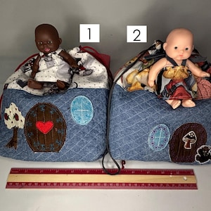 May include: Two handmade doll bags with denim-style fabric and decorative appliqués. Each bag features a doll on top. One bag has a tree and heart design, the other has a mushroom and bear design. A ruler is at the bottom.
