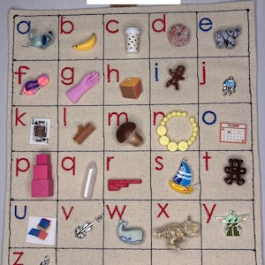 May include: A beige fabric alphabet chart labeled Set F1 with black grid lines and embroidered letters from a to z. Each square contains a small miniature object or charm representing the corresponding letter, including a banana, donut, elephant, and sailboat.