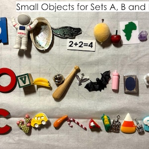 May include: Assorted small objects, including a blue letter "a", a toy astronaut, a seashell, a green bat wing, a yellow fruit, a red apple, a map of Africa, a purple gem, a green block, a banana, a baseball bat, a silver bell, a black bat, a pink baby bottle, a book, a white bird, a cactus, a yellow car, a carrot, a candy cane, a slice of cake, a corn, a castle, a candy corn, a crown, and a cupcake. The text "Small Objects for Sets A, B and E" and "2+2=4" are also visible.