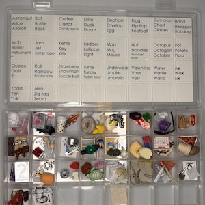 May include: A clear plastic storage container with a grid of compartments containing various miniature objects. The lid features a printed list of words organized alphabetically from A to Z, with corresponding small items stored in the sections below.