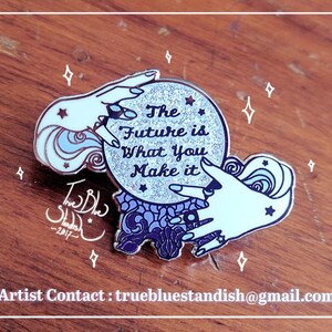 May include: A metal enamel pin featuring a glittery silver crystal ball held by two pale hands. The crystal ball displays the text The Future is What You Make it. The base is dark purple with ornate patterns, and small stars surround the design.