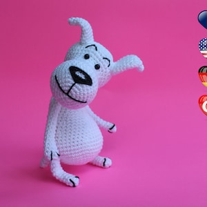 Könnte beinhalten: Weißes gehäkeltes Hundespielzeug mit schwarzen Augen, Nase und Mund. Das Spielzeug sitzt auf einem rosa Hintergrund. Im Hintergrund befinden sich vier Herzen mit Flaggen verschiedener Länder.