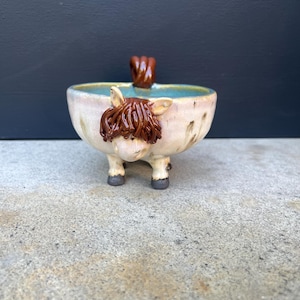 May include: A ceramic bowl shaped like a Highland cow, featuring a light cream-colored body with brown glazed hair, ears, and a tail. The interior of the bowl is glazed in a soft blue, and the base is supported by four small, dark-colored hooves.