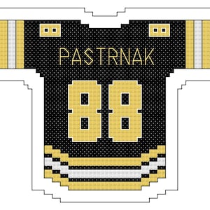 Op de afbeelding: Zwart en goud hockey shirt met de naam "PASTRNAK" en het nummer "88" in goud. Het shirt heeft gouden en witte strepen op de mouwen en zoom. Het ontwerp is een gestileerde, gepixelde weergave.