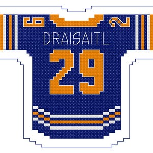 Op de afbeelding: Kruissteekontwerp van een blauw en oranje hockeytrui. De trui heeft de naam "DRAISAITL" en het nummer "29" in oranje. De trui heeft oranje en witte strepen op de mouwen en onderkant.