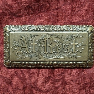 May include: A gold-colored metal plaque with an ornate border and the words "At Rest" in a decorative font.