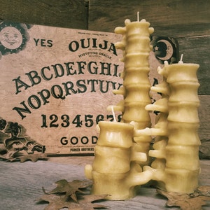 Pu&ograve; includere: Un set di quattro candele di cera d'api gialle a forma di colonna vertebrale. Le candele sono disposte a forma di torre e sono posizionate su una superficie di legno davanti a una tavola Ouija vintage.