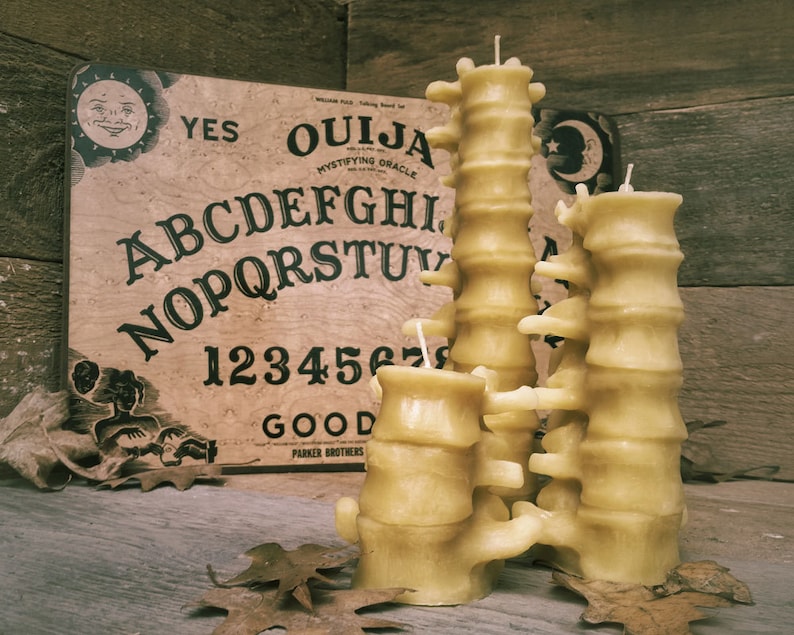 Pu&ograve; includere: Un set di quattro candele di cera d'api gialle a forma di colonna vertebrale. Le candele sono disposte a forma di torre e sono posizionate su una superficie di legno davanti a una tavola Ouija vintage.