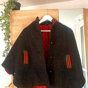 May include: A dark charcoal gray cape featuring a vibrant red interior lining with a repeating floral pattern. The garment includes a high collar, vertical slit pockets accented with red and green trim, and metallic buttons along the front closure.