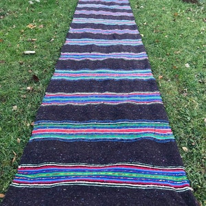 Puede incluir: Una alfombra larga, tejida a rayas negras y multicolores con flecos en los extremos. La alfombra está colocada sobre un césped.