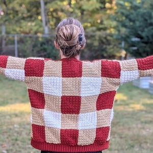 Può includere: Un maglione fatto a mano con un motivo a scacchiera rosso, beige e bianco. Il maglione ha maniche lunghe e un orlo a coste. La persona è in piedi all'aperto con le braccia tese, mostrando il design del maglione.