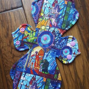 May include: A reusable cloth menstrual pad featuring a colorful stained-glass design with images of a castle, characters, and a rose. The pad has a cross shape with snap closures and a blue, purple, and yellow color scheme.