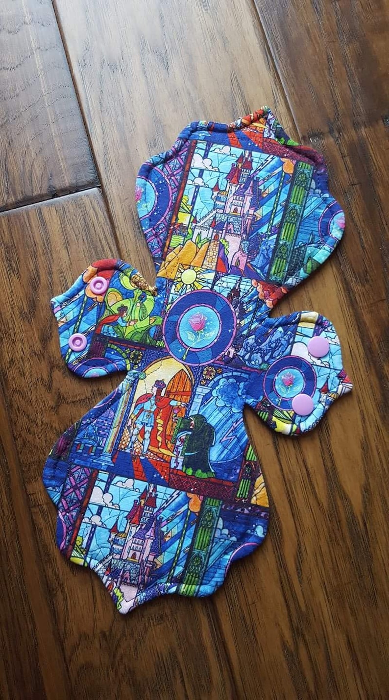 May include: A reusable cloth menstrual pad featuring a colorful stained-glass design with images of a castle, characters, and a rose. The pad has a cross shape with snap closures and a blue, purple, and yellow color scheme.